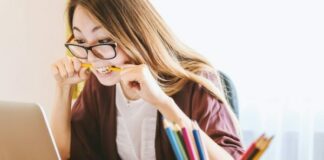 woman biting pencil while sitting on chair in front of computer during daytime
